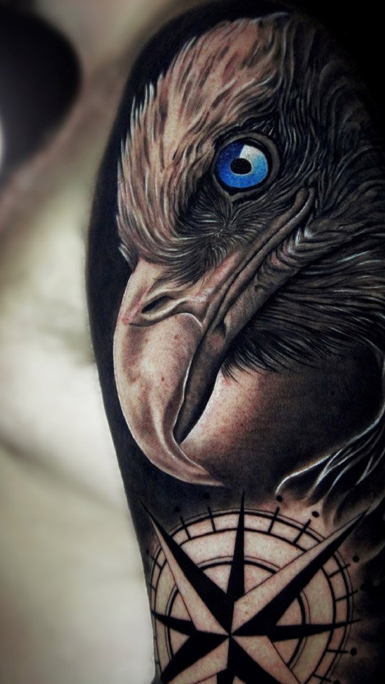 Tatuagem realista de águia com olho azul sobre rosa dos ventos, em preto e cinza